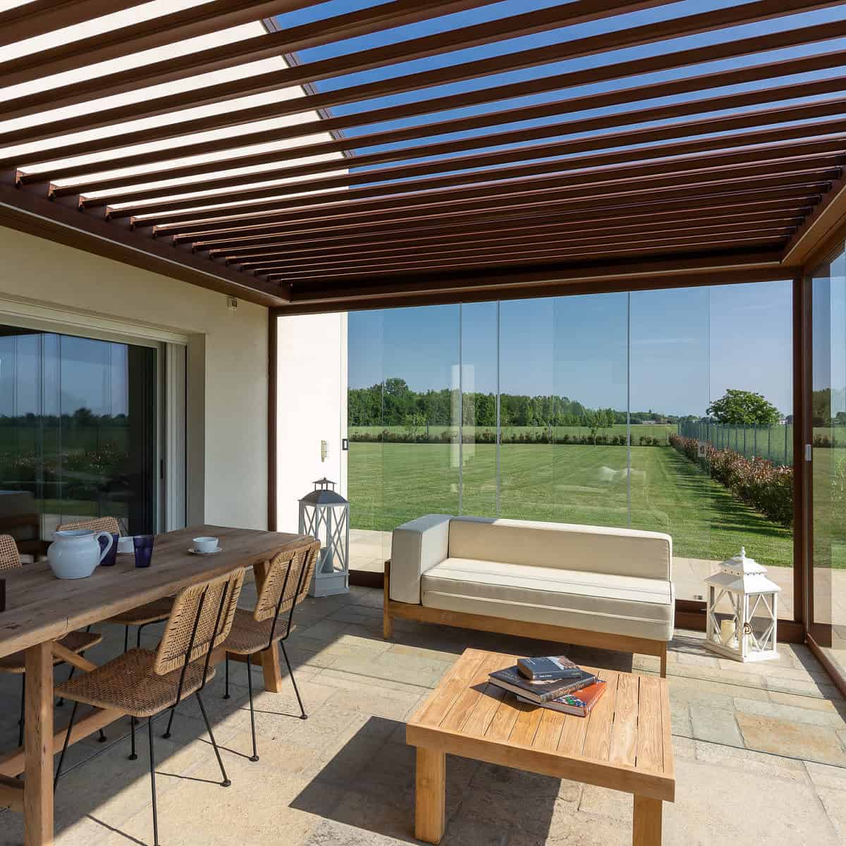 This image showcases the inside view of a Kedry Prime louvred roof pergola in brown, equipped with rotating louvres and glass sliding doors. With the louvres adjusted to a partially open position, natural light fills the space while still providing shade and shelter from the elements. The glass sliding doors offer a seamless connection between the indoor and outdoor areas, making it a perfect space for entertaining, dining, or simply relaxing. The versatile and stylish design of the Kedry Prime louvred roof pergola makes it a valuable addition to any home, providing an attractive and functional outdoor living space that can be used year-round.