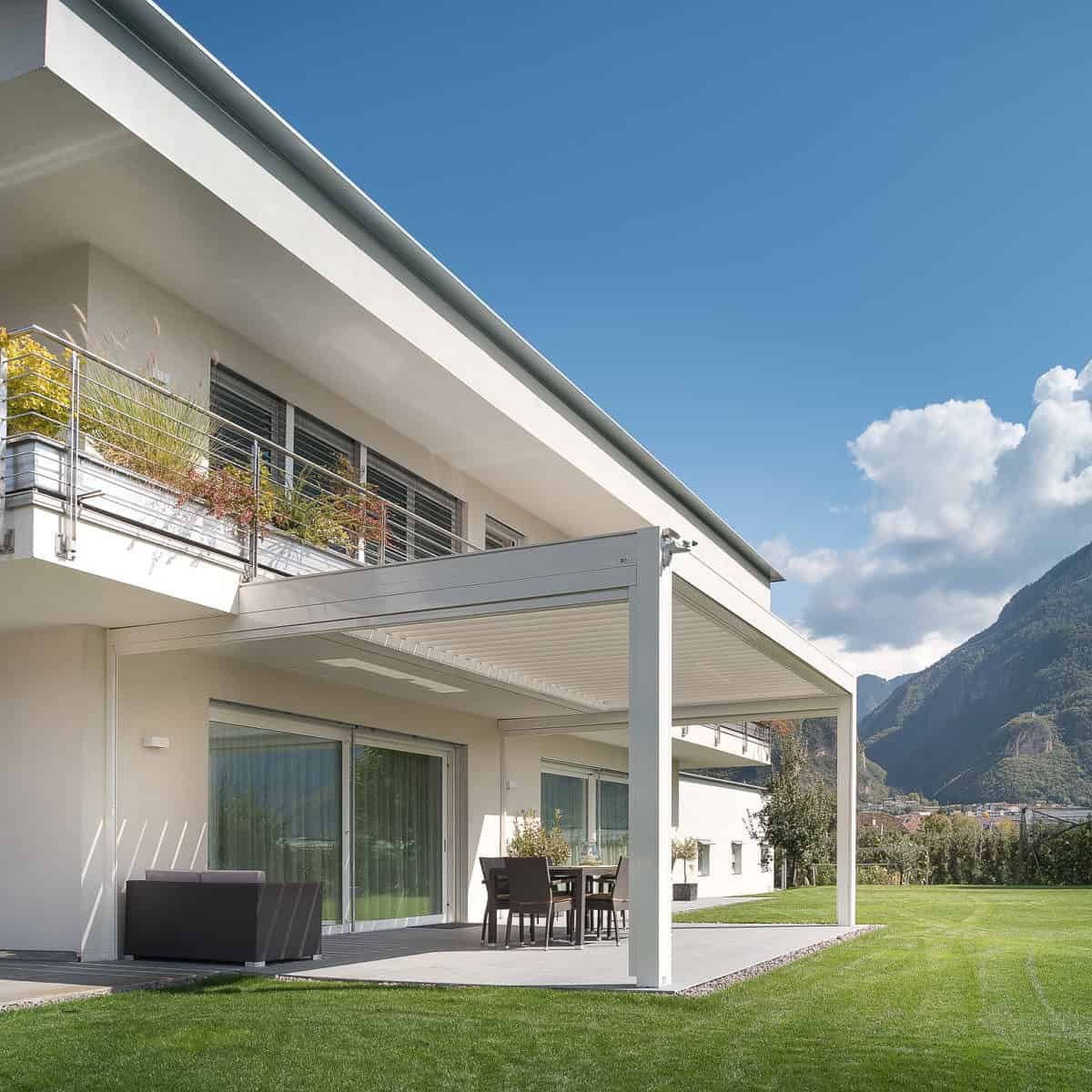This image showcases a KE Kedry Prime louvred roof pergola in white, wall mounted over a patio area. With its sleek and stylish design, the Kedry Prime pergola not only adds an attractive element to the outdoor space but also provides practical benefits with its adjustable louvres, allowing you to control the amount of sunlight and shade. The white color of the pergola blends seamlessly with the patio and surrounding landscaping, creating a unified and cohesive outdoor living space. The wall-mounted design maximizes space efficiency and ensures the pergola doesn't take up valuable floor space on the patio. With the KE Kedry Prime louvred roof pergola, you can enjoy your outdoor space in comfort and style, no matter the weather.