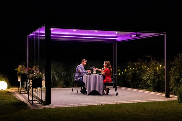 Couple sipping wine under a louvred roof pergola illuminated by ambient LED lights.
