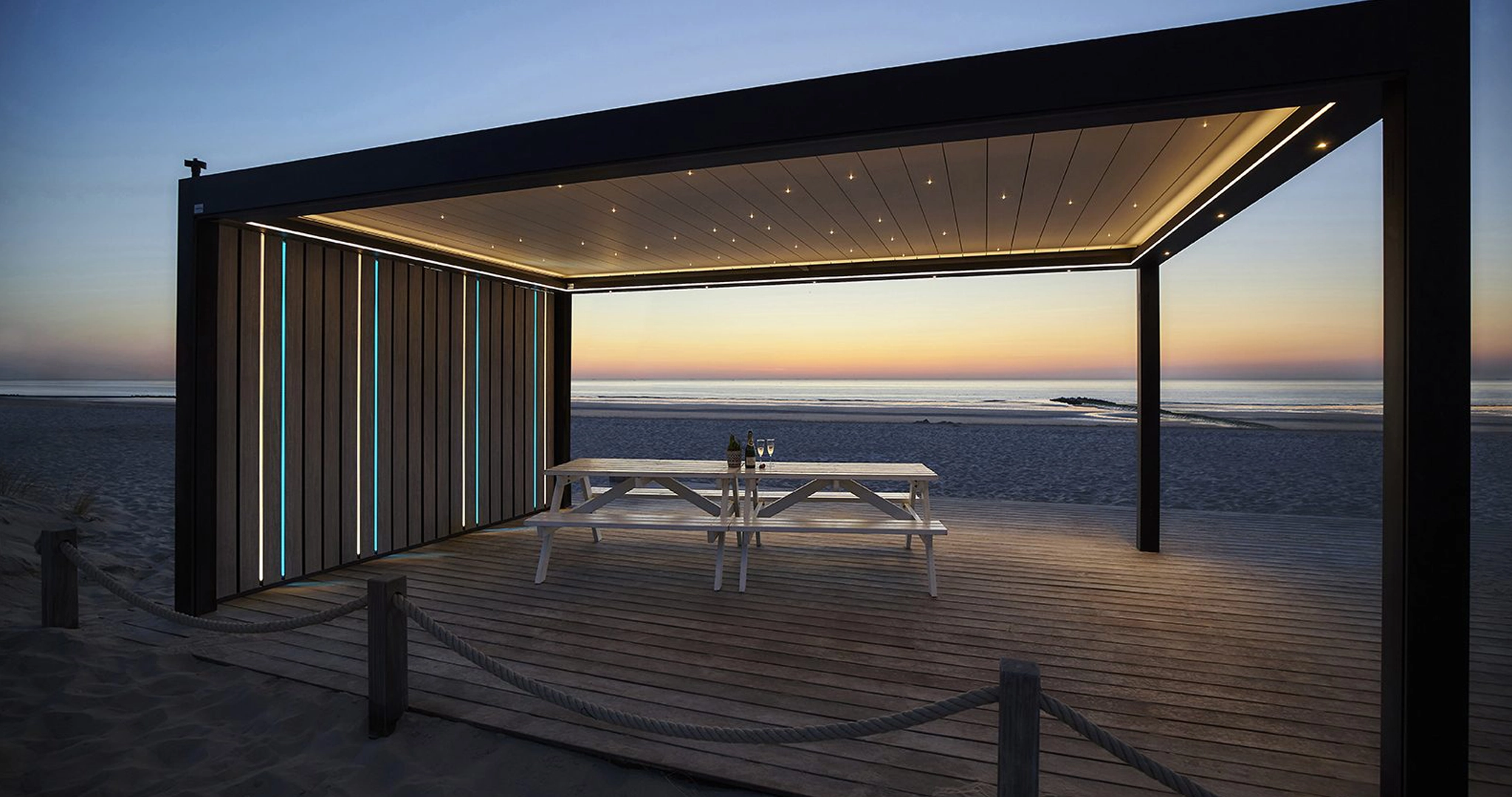 Beachside louvred roof pergola illuminated by LED lights, with a stylish wooden panel wall.